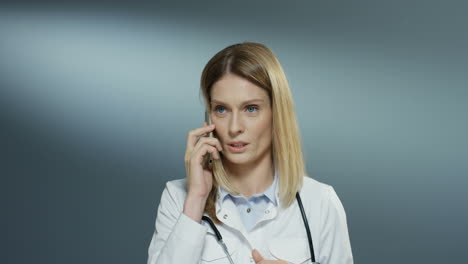 Good-looking-young-Caucasian-female-physician-in-white-medical-gown-speaking-on-the-mobile-phone-like-solving-some-problems-and-explaining-issues.